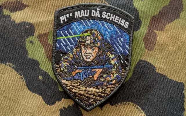 Zugbadge ricamato "FI* MAU DA SCHEISS" con soldato in mimetica sotto la pioggia, tema tiro notturno.