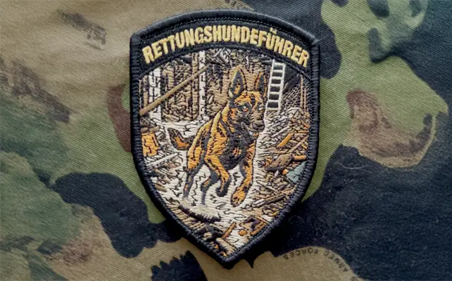 Zugbadge ricamato "RETTUNGSHUNDEFÜHRER" con cane da soccorso tra le macerie, motivo ricerca e salvataggio in stile svizzero.
