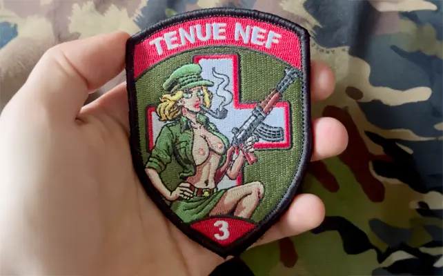 Zugbadge ricamato "TENUE NEF" con pin-up soldatessa, fucile e croce sanitaria sullo sfondo, stile militare svizzero.